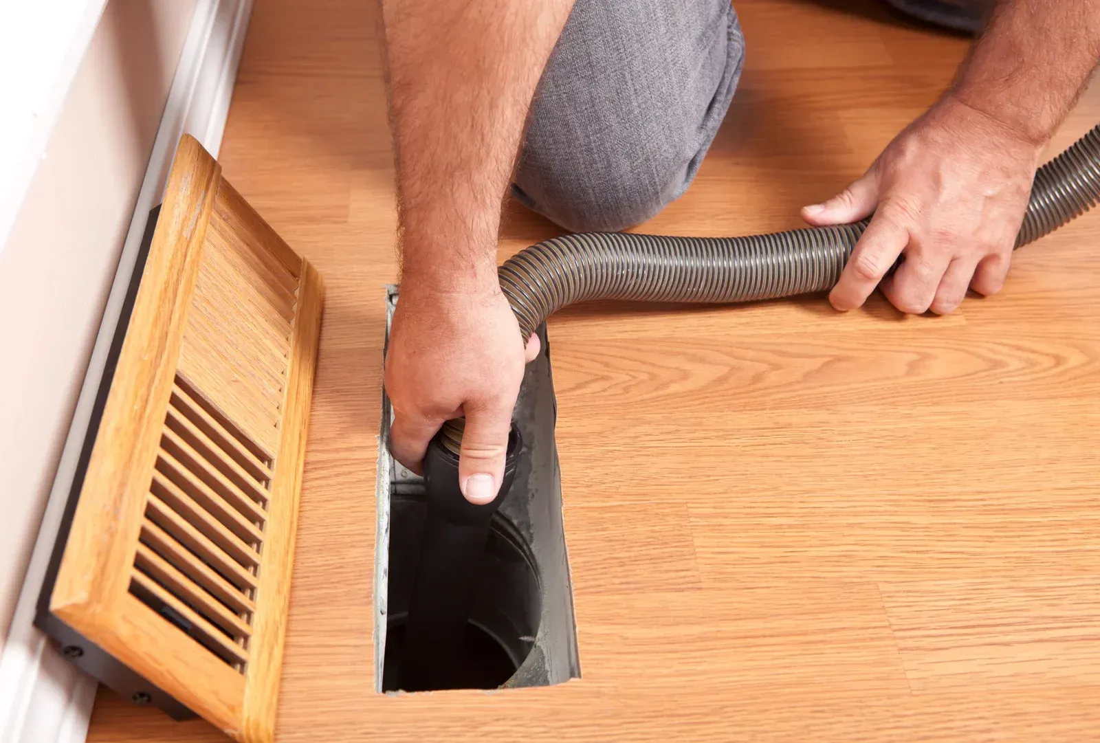 Person using a vacuum hose to clean a floor vent on a wooden floor