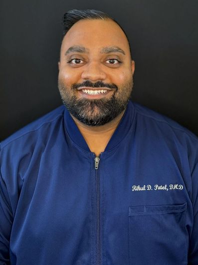 A man with a beard smiles, wearing a blue jacket against a black background.