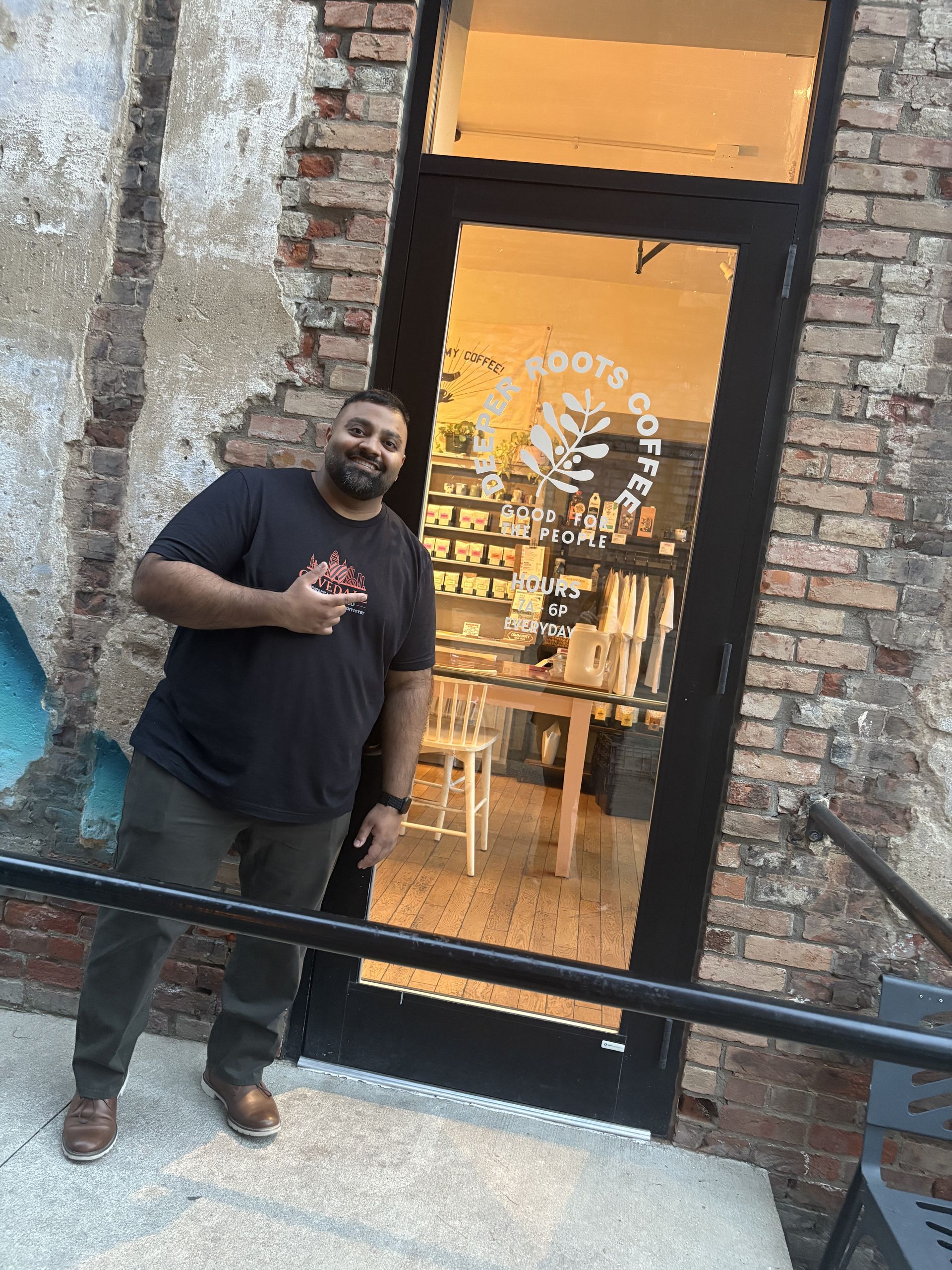 Man smiling, pointing in front of Roots Coffee shop, brick exterior.