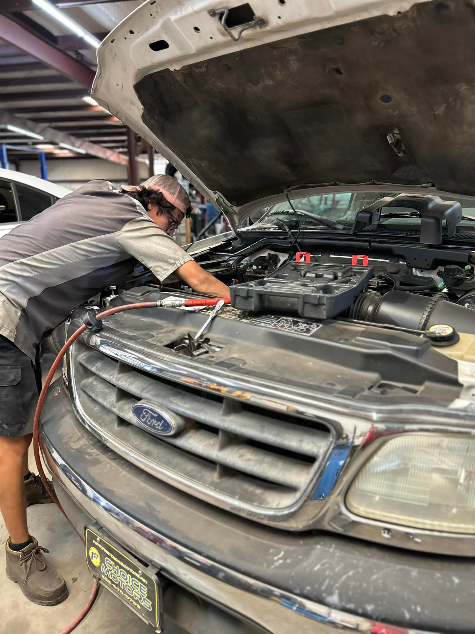 Mechanic working on the engine of a Ford truck with the hood open, holding jumper cables in a garage.