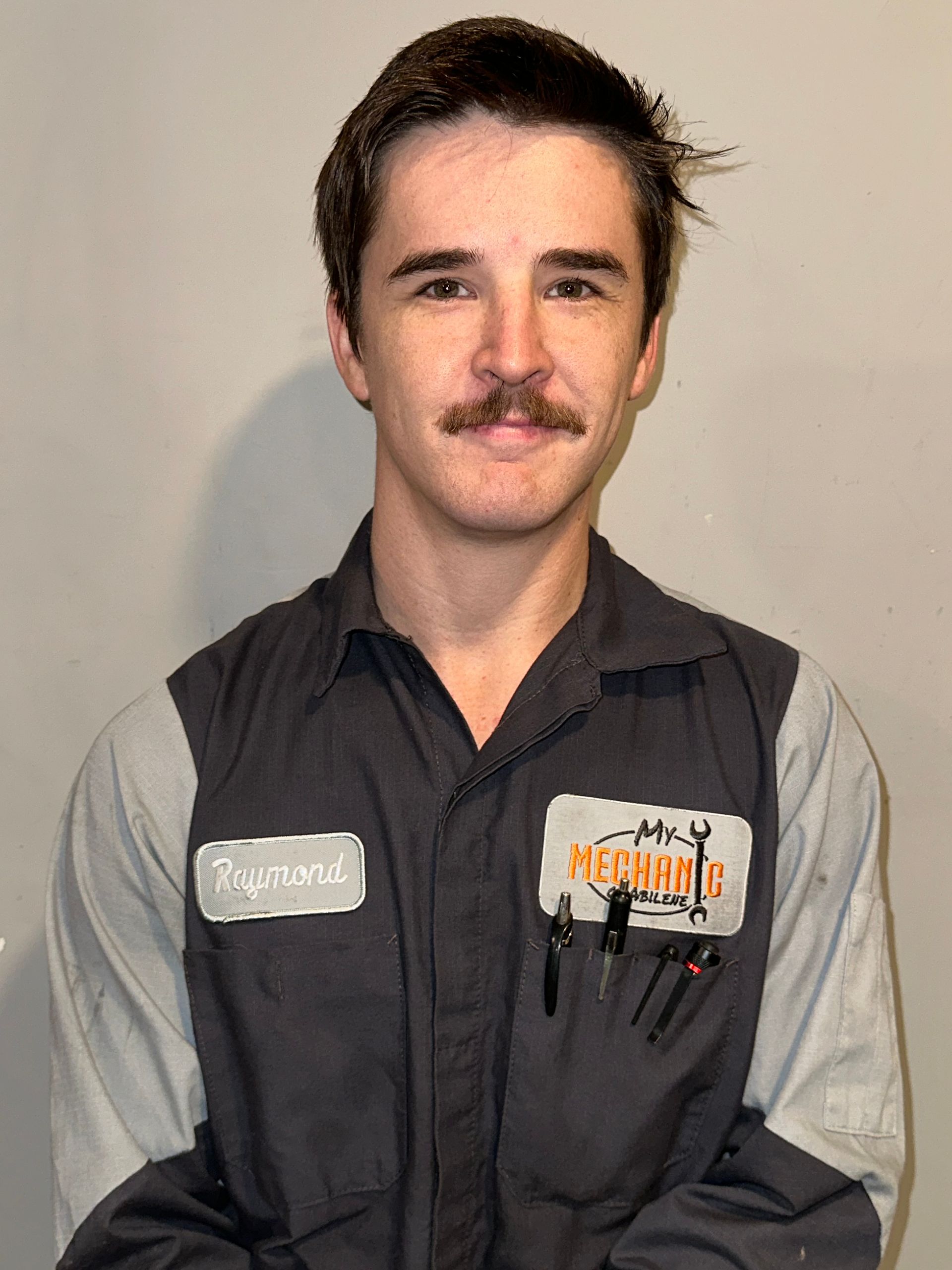 Mechanic with crossed arms in auto shop, wearing work uniform, tattoos visible.