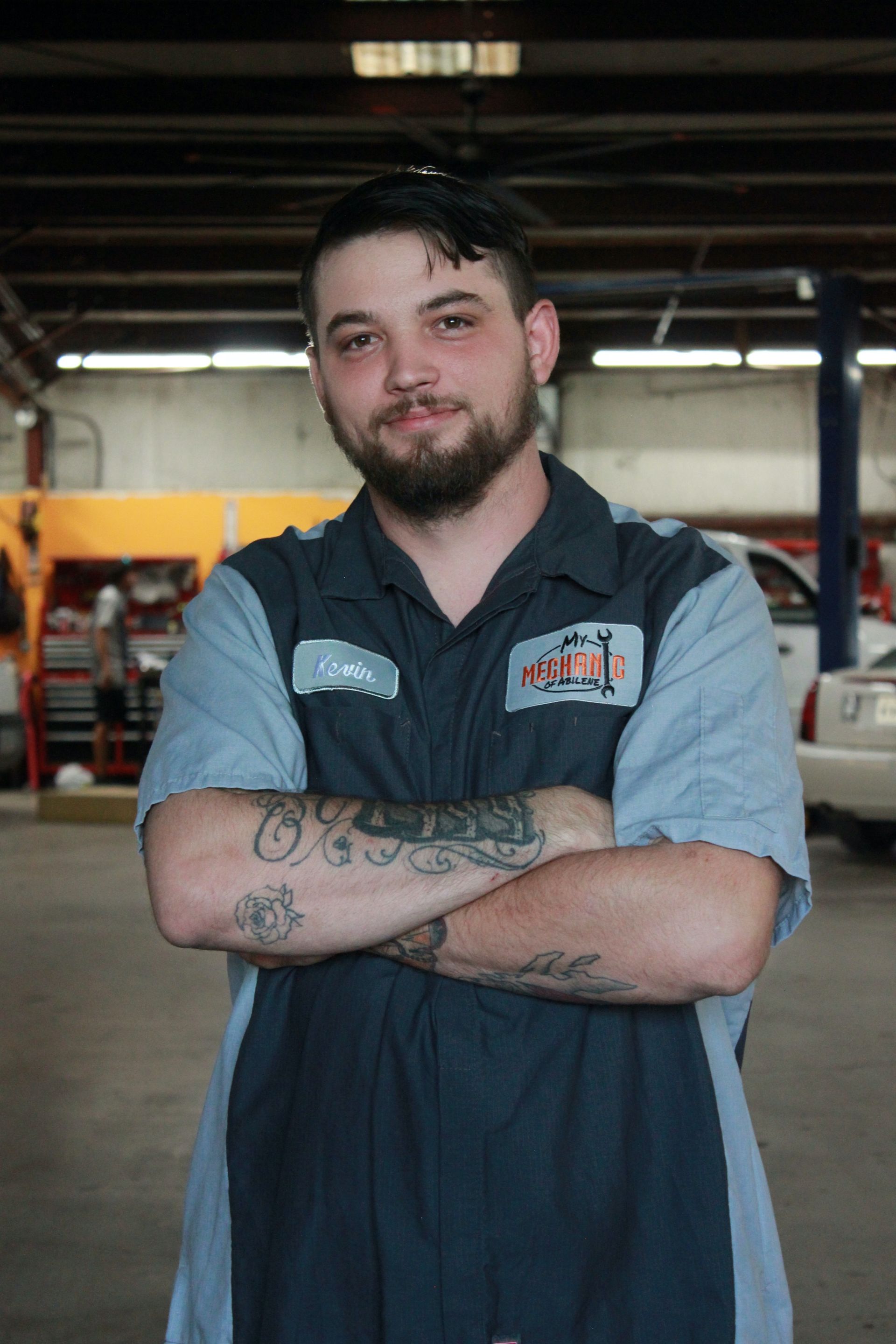 Mechanic with crossed arms in auto shop, wearing work uniform, tattoos visible.