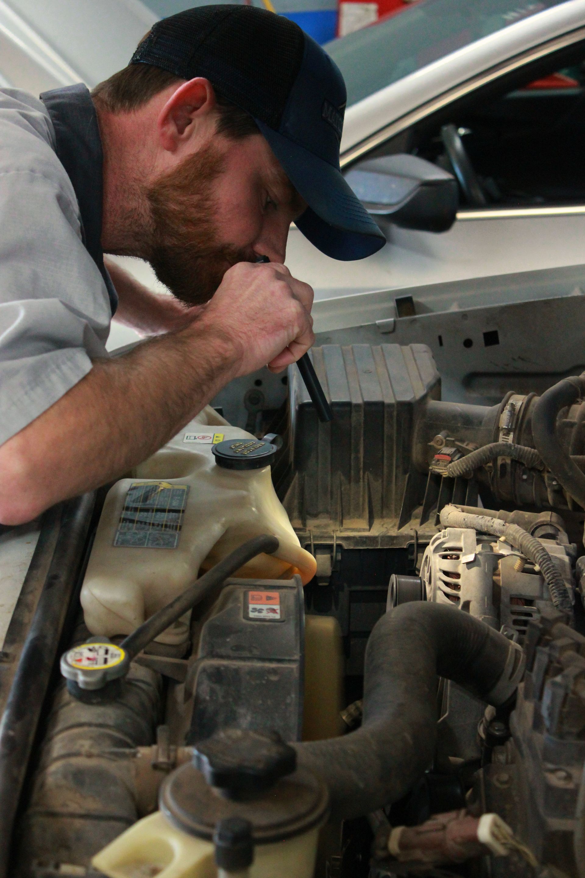 Mechanic inspecting engine compartment with a flashlight. He's wearing a blue cap, working inside a garage.