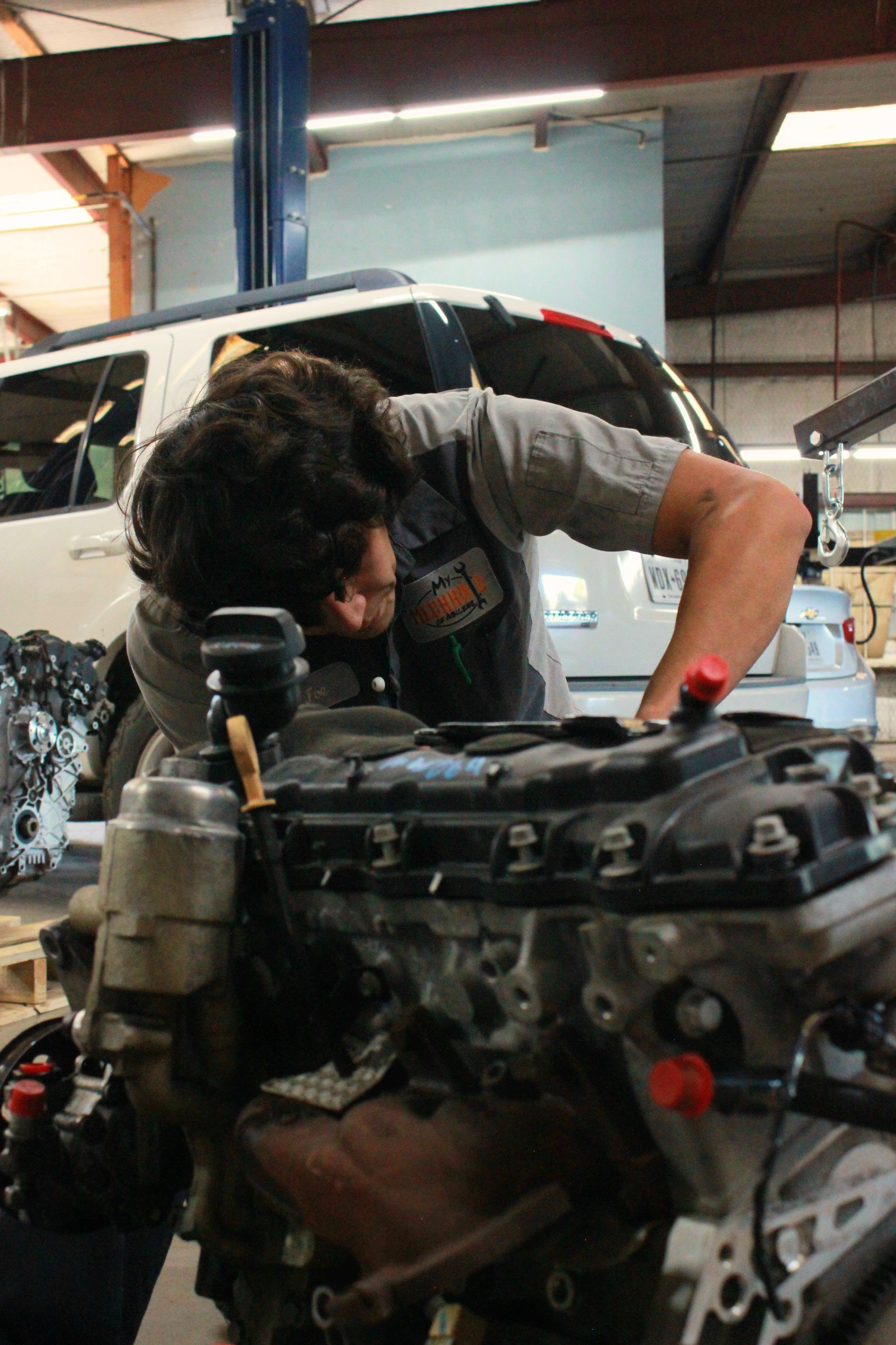 Mechanic working on an engine in a garage. Wearing gray shirt, looking down, hands on engine. Vehicle in background.