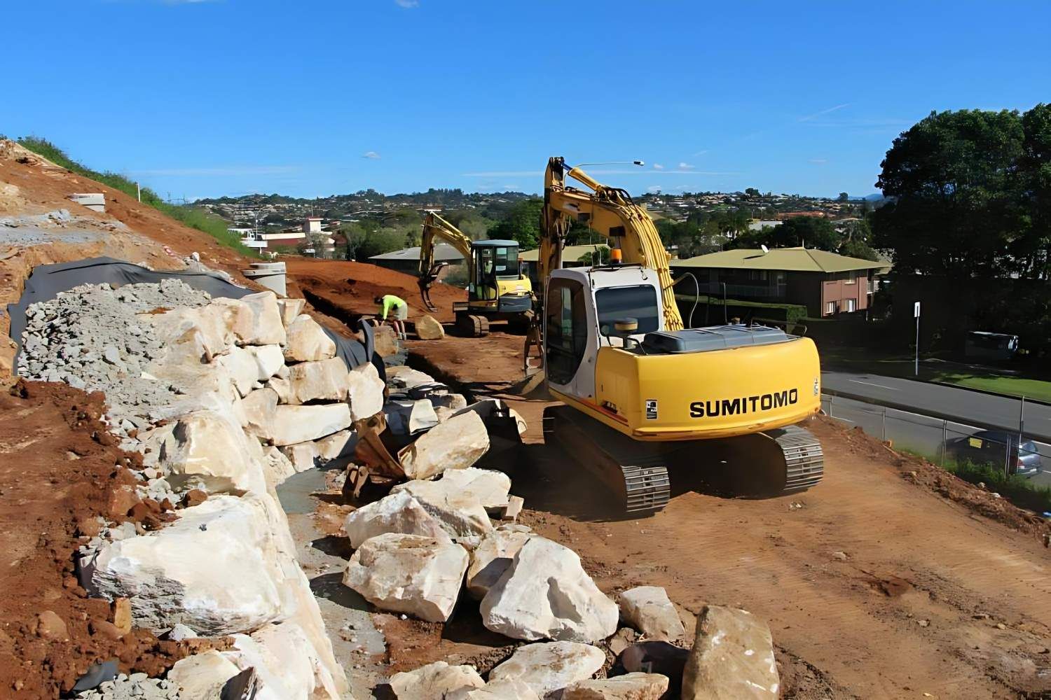 Excavator placing large sandstone blocks while building a retaining wall on a Gold Coast hillside property, with the suburb skyline in the background
