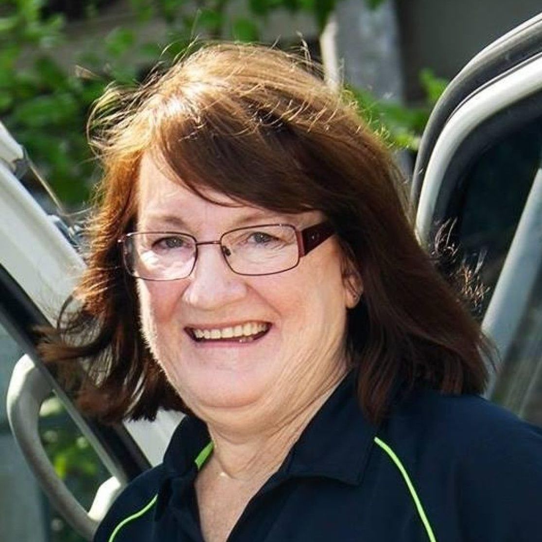 Woman with brown hair and glasses, smiling, in a dark shirt, near a vehicle.