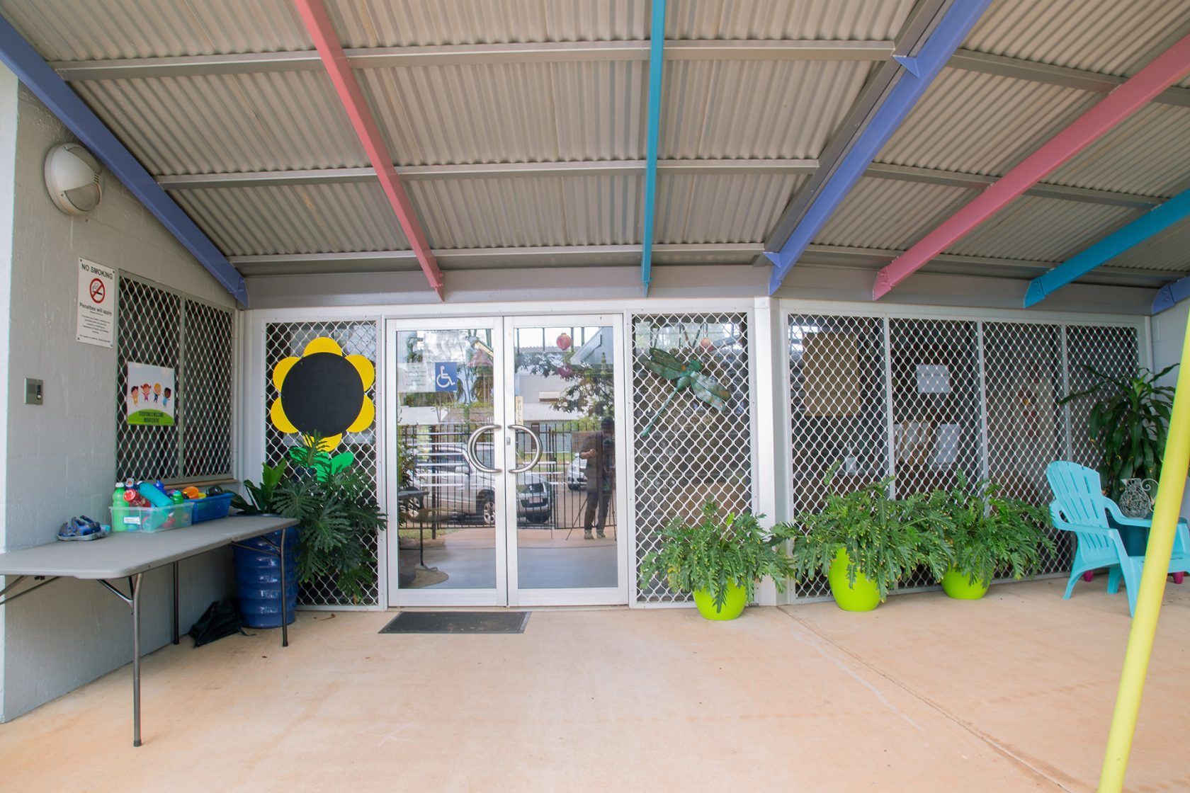 Entrance to a building with glass doors, wire screens, colorful beams, and potted plants.