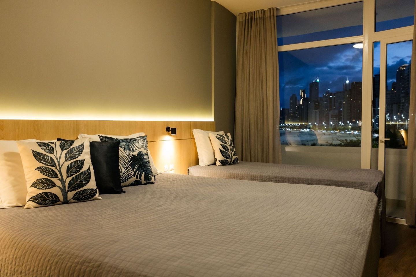 Quarto de hotel com duas camas bem arrumadas, iluminação aconchegante e vista para o horizonte da cidade através da janela.