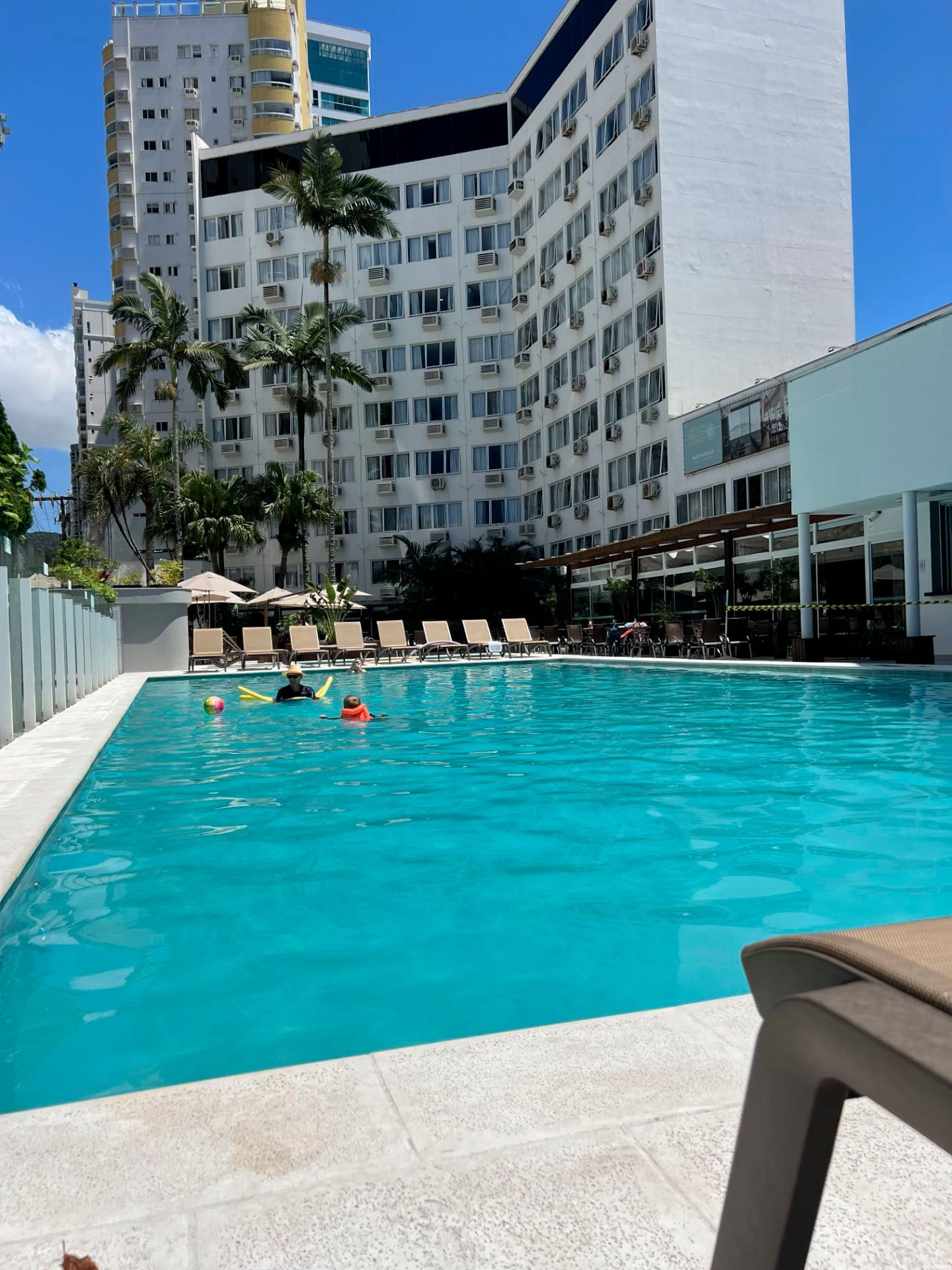 Piscina de hotel com água turquesa, espreguiçadeiras e um edifício branco curvo sob um céu azul límpido.