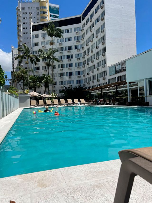 Piscina de hotel com água turquesa, espreguiçadeiras e um edifício branco curvo sob um céu azul límpido.