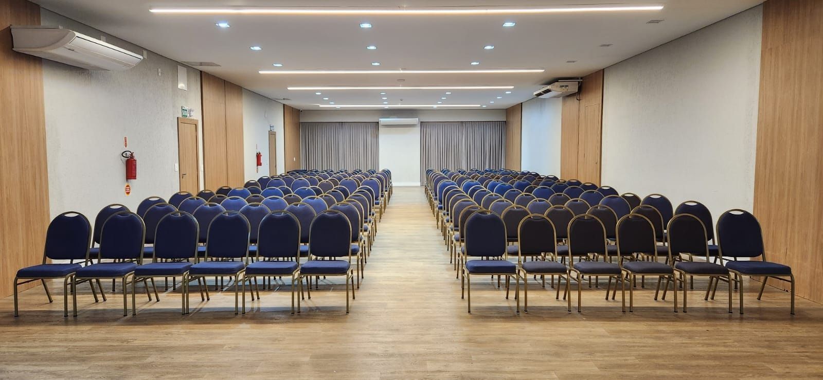 Sala de conferências vazia com fileiras de cadeiras azuis voltadas para uma parede frontal com cortinas.