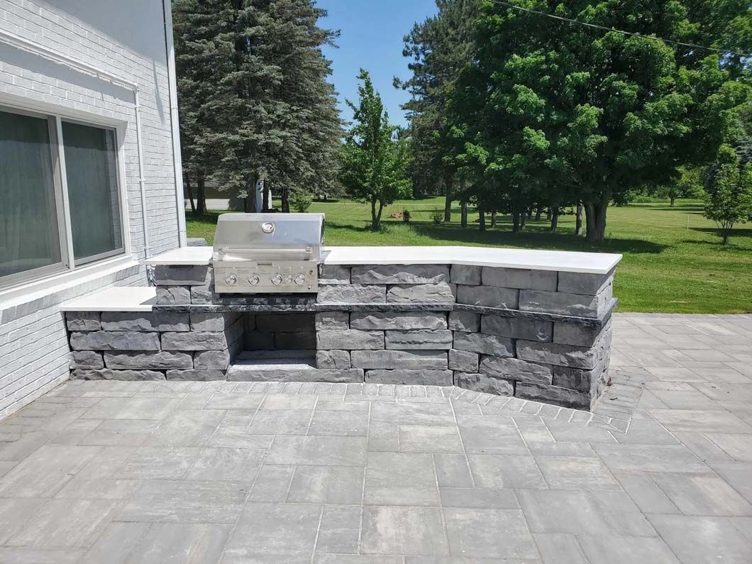 Stone outdoor kitchen with grill on a patio beside a house and green lawn