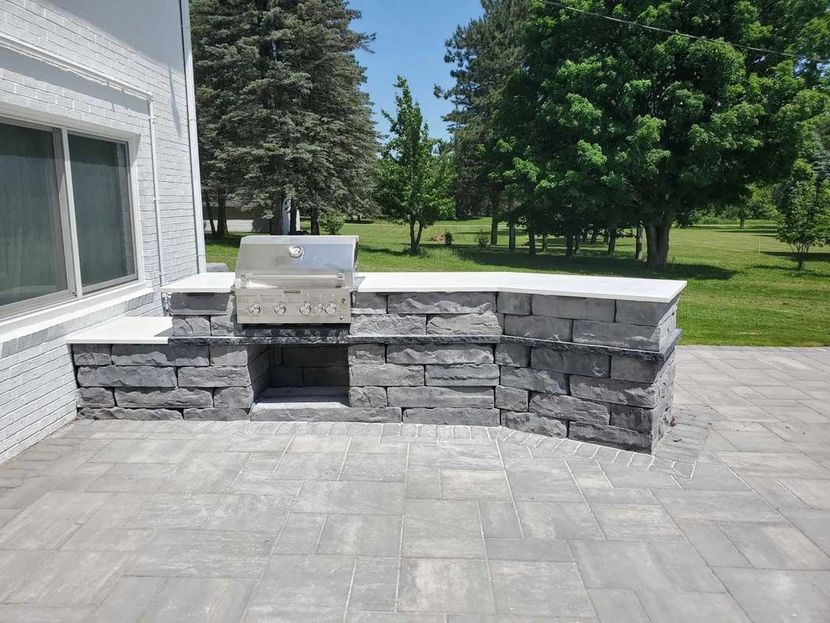 Stone outdoor kitchen with grill on a patio beside a house and green lawn