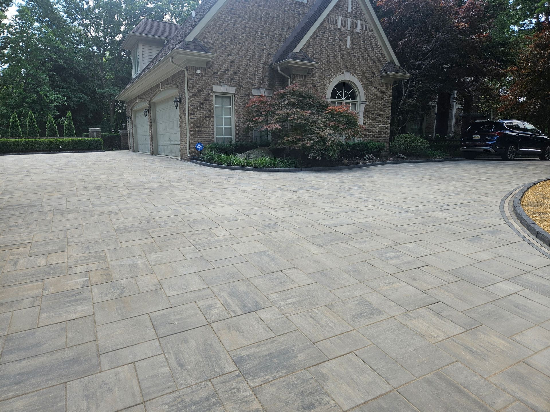 Large stone-paved driveway beside a brick house with garages and landscaping, with a dark car parked at right.