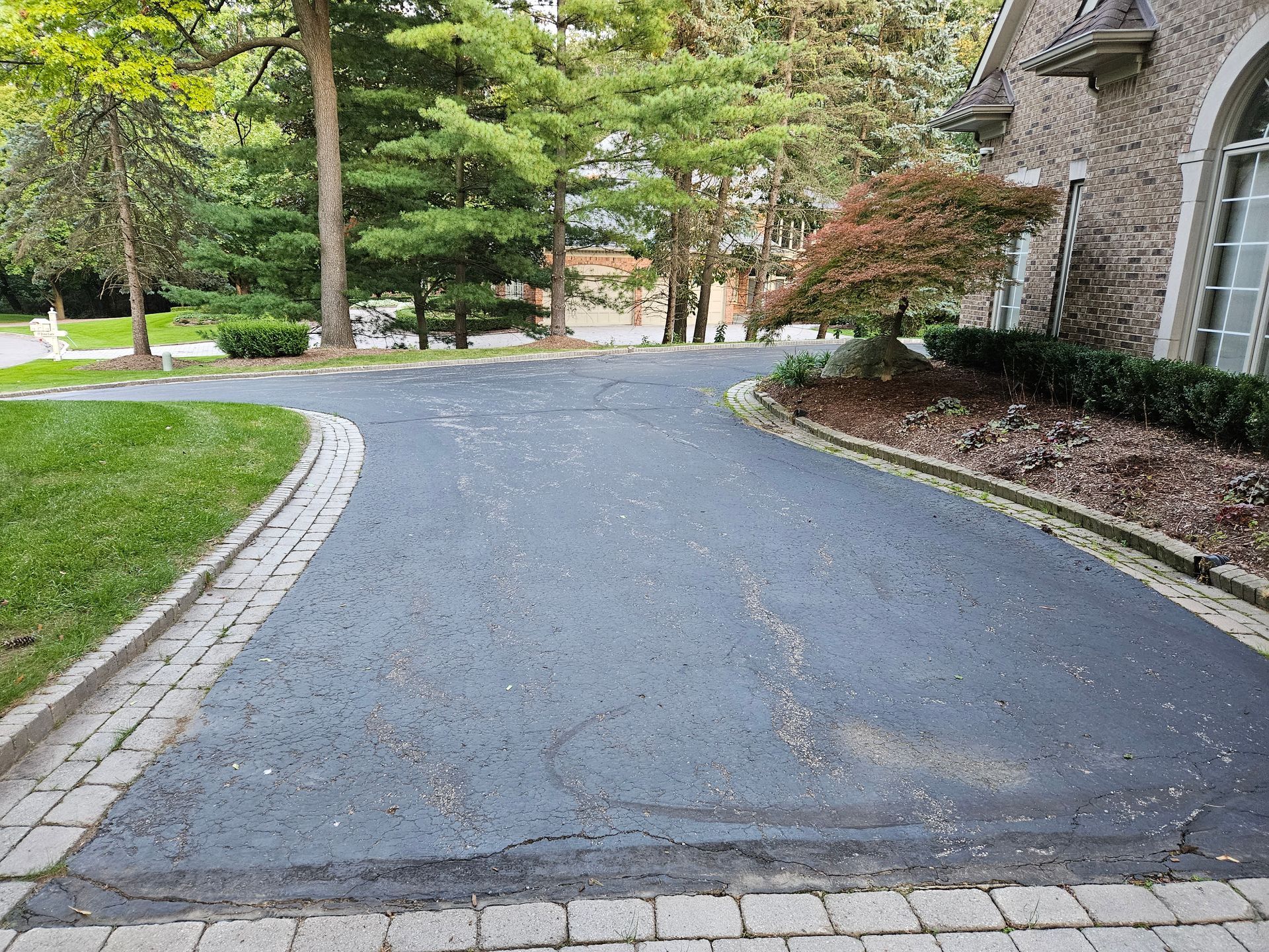 Curved driveway beside a stone house, bordered by grass and trees on a quiet residential street