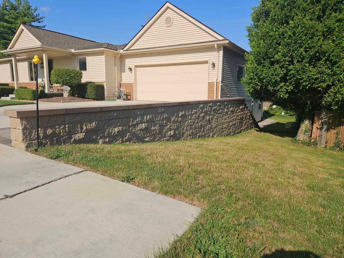 Suburban house with beige siding, garage, front lawn, and a sloped driveway beside a stone retaining wall