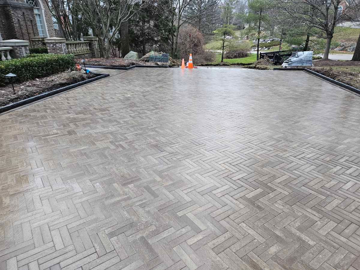 Rainy brick-paved driveway leading to a landscaped yard with trees and parked cars in the distance