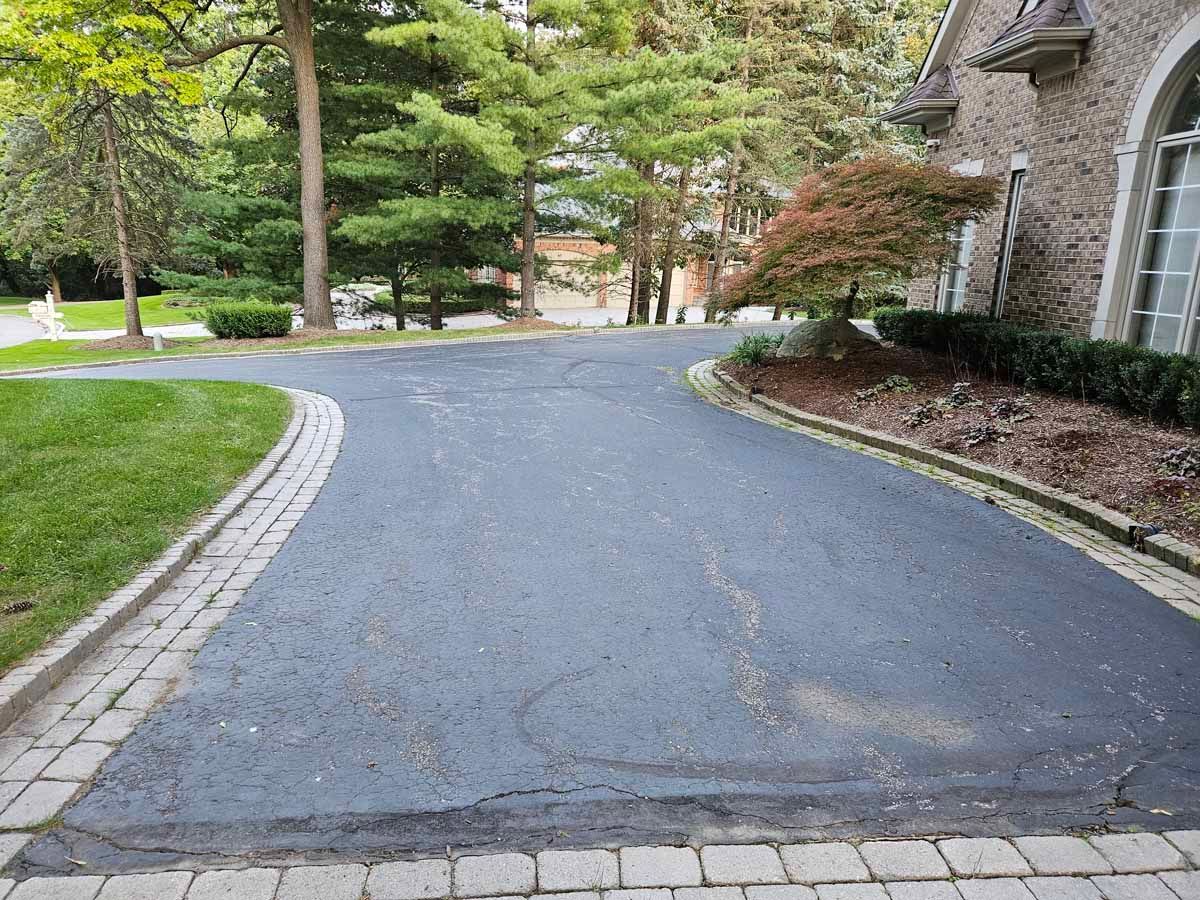 Curved asphalt driveway beside a stone house, lined with trees and green lawn