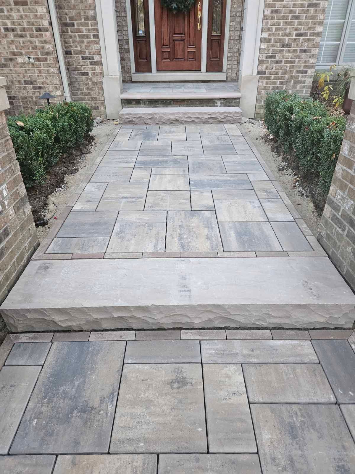 Front walkway with gray stone pavers, a step, and a wooden front door between brick walls and shrubs