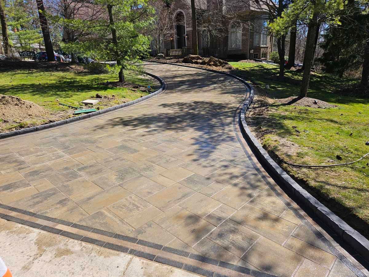 Curved brick walkway leading through a landscaped garden with trees and grass in sunlight