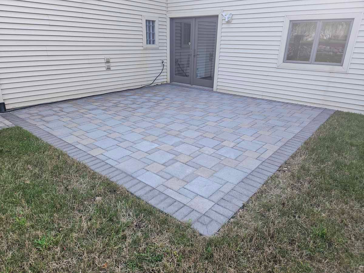 Paver patio beside a house with a sliding glass door, bordered by grass.