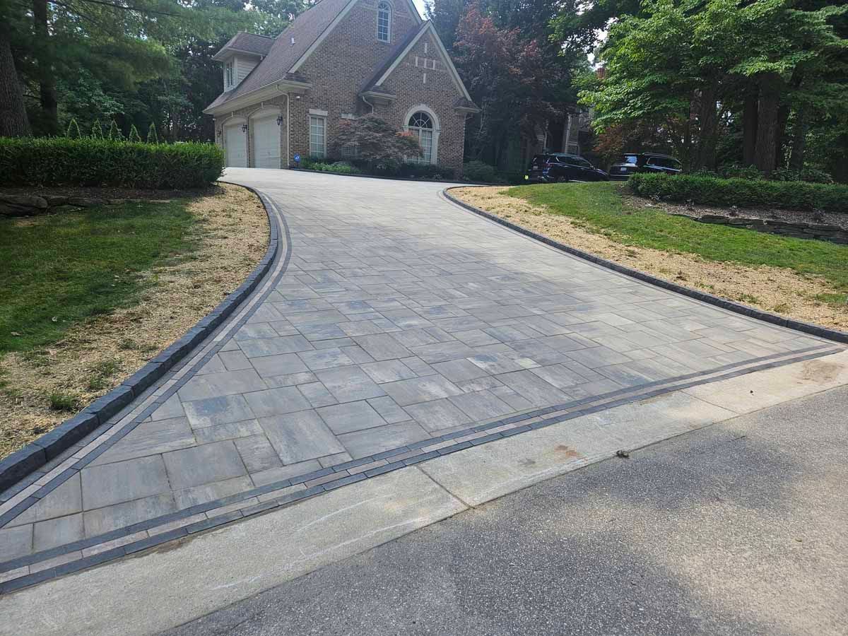 Stone-paved driveway curving to a house with a garage, bordered by manicured hedges and trees