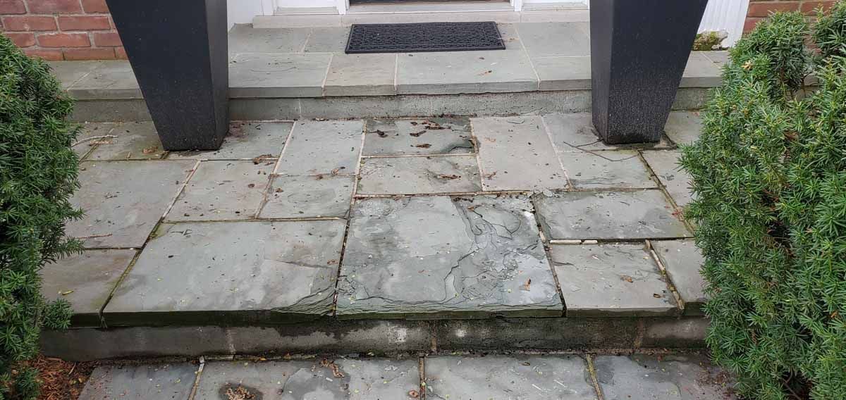 Broken stone walkway leading to a front porch, with uneven slabs and steps between shrubs