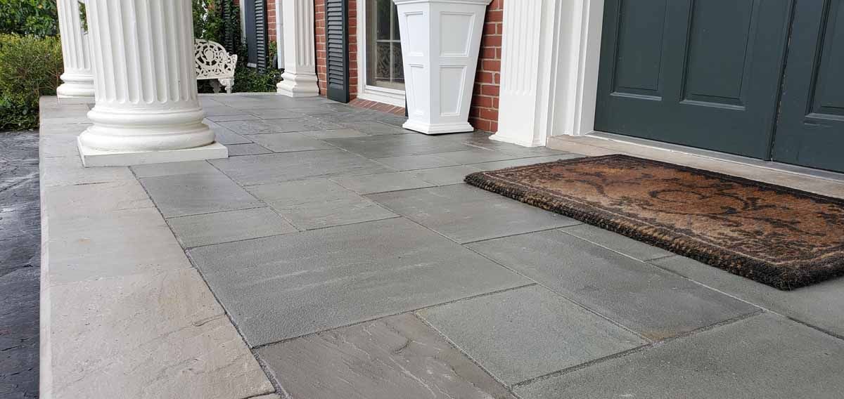 Stone walkway with white columns and dark doors beside a brown doormat