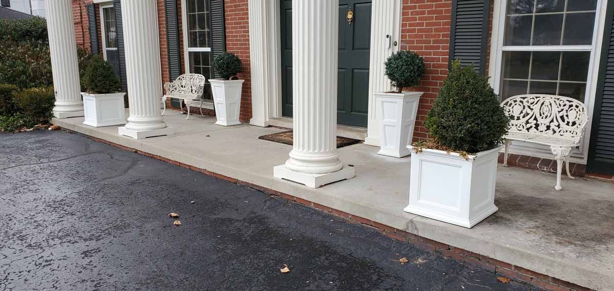 White columns and potted topiaries line a porch beside a wet driveway and wooden bench.