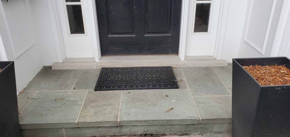 Front porch with stone steps, black doormat, and two square planters by a white door