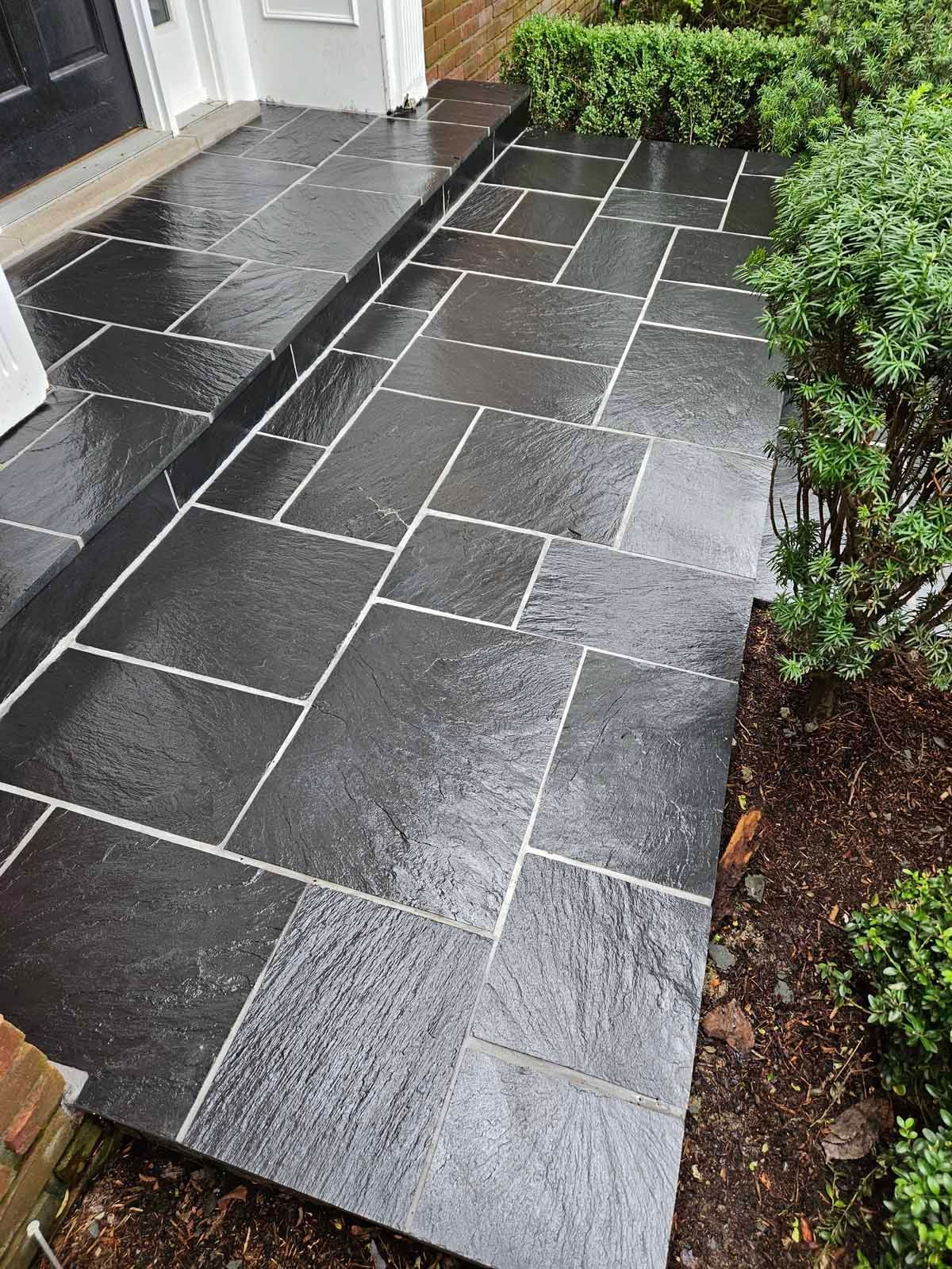 Wet black stone walkway leading to a front door, bordered by white walls and green shrubs.