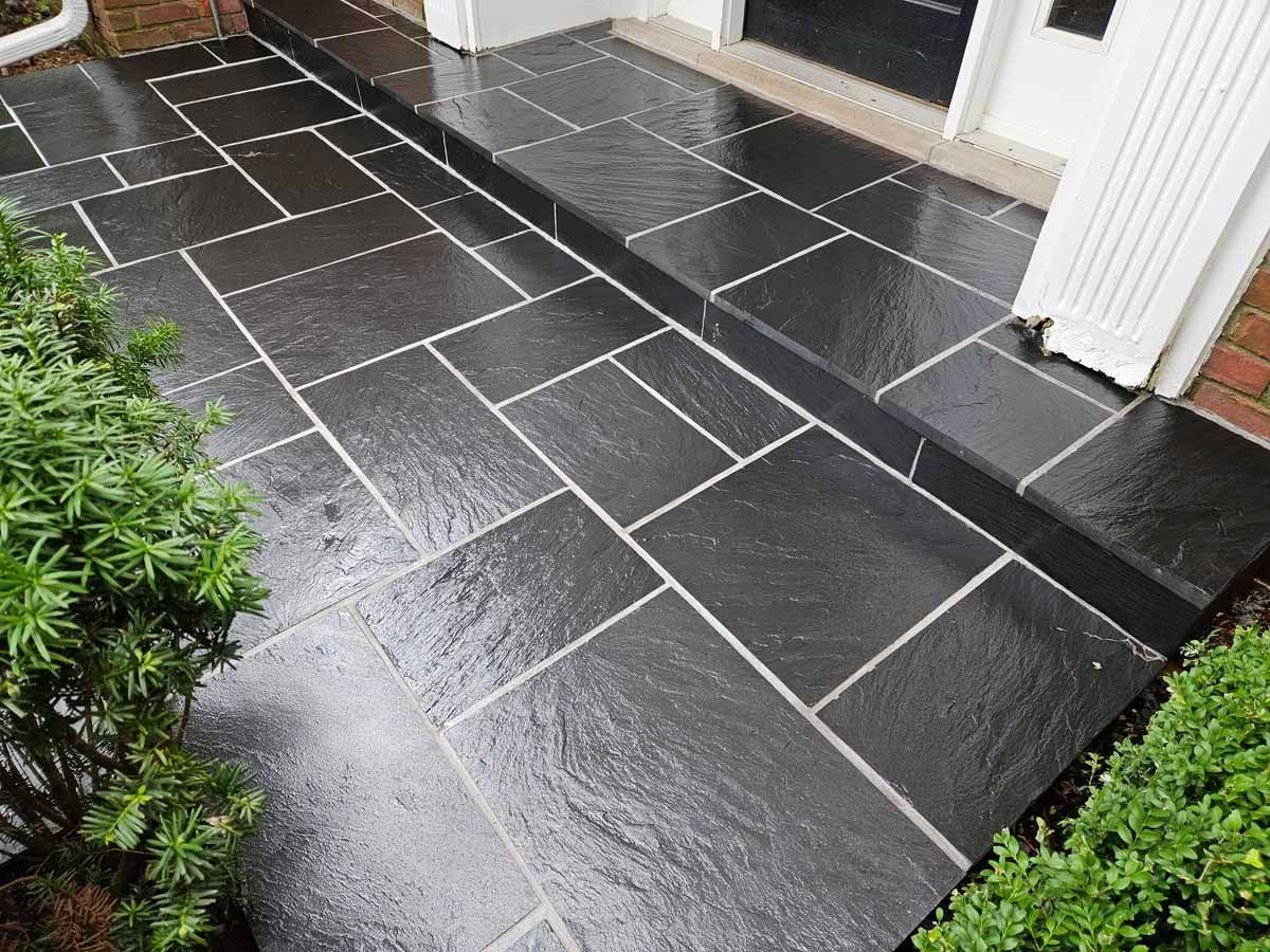 Wet dark stone patio tiles by a house entrance, with a step and shrub at the edge