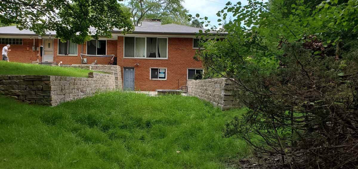 Brick house with large windows behind a stone retaining wall and overgrown green lawn