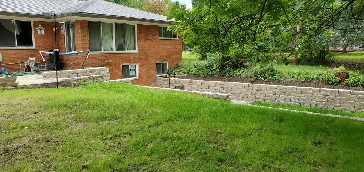 Red brick house beside a grassy yard and stone retaining wall with trees in the background