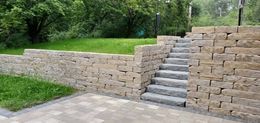 Stone steps in a landscaped retaining wall beside a paved path and green grass.