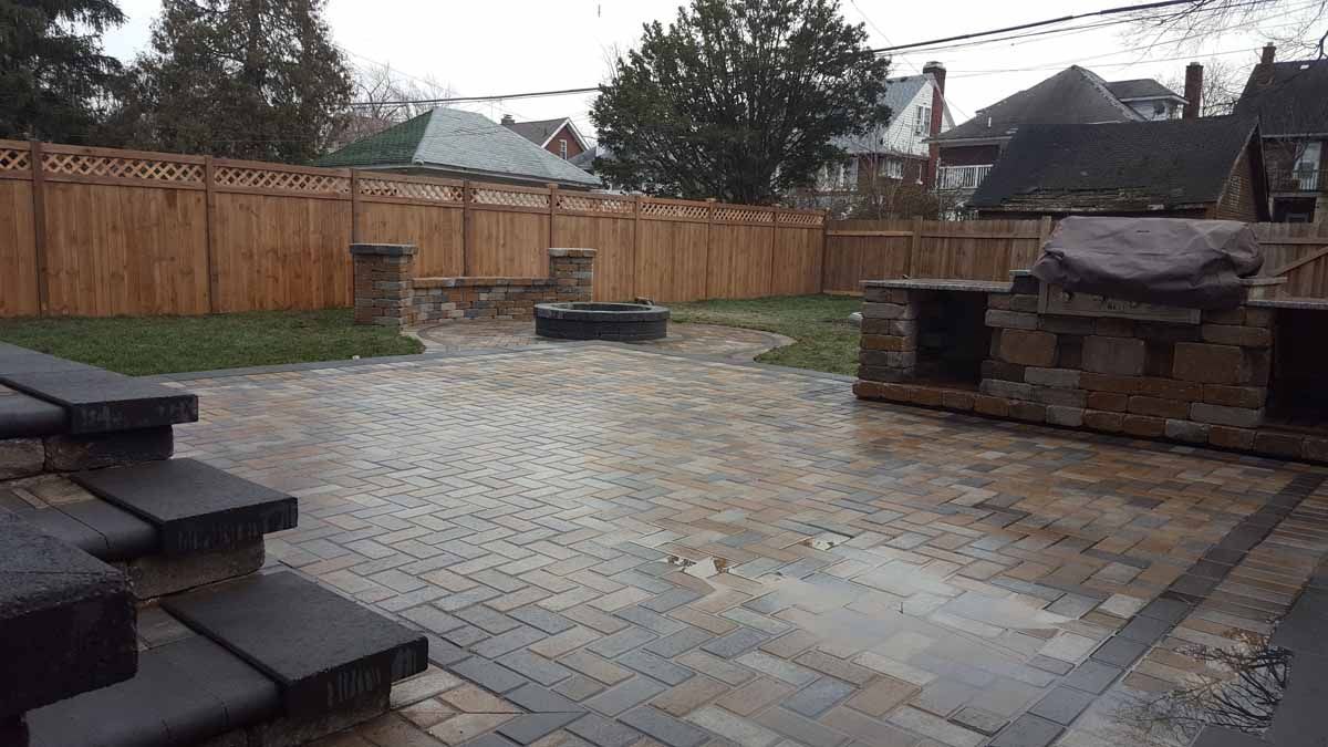 Rainy backyard patio with wet stone paving, wooden fence, and stone steps and grill in the corner