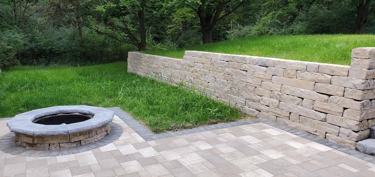 Stone fire pit beside a paved patio and tan retaining wall in a grassy backyard
