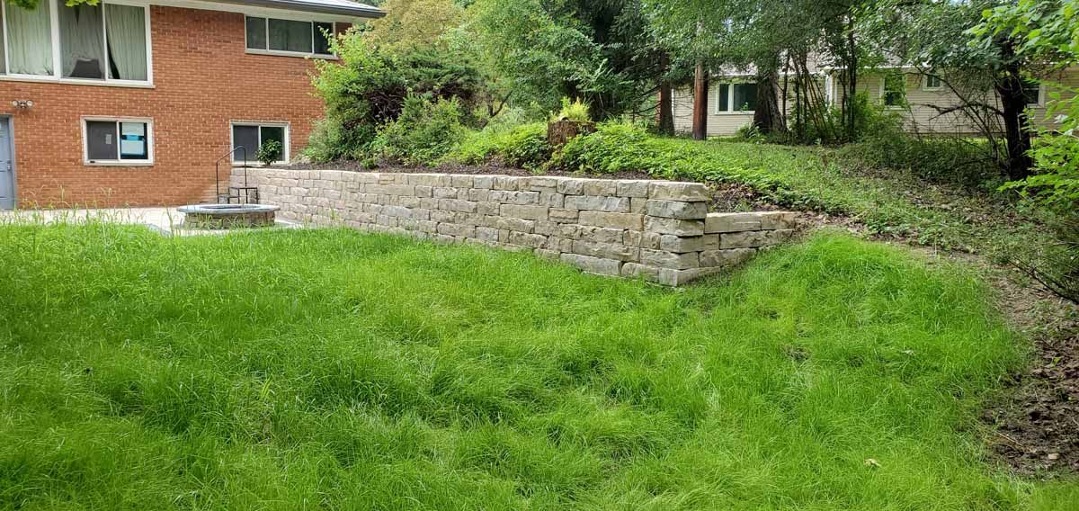 Grassy backyard with a stone retaining wall beside a red brick house and trees