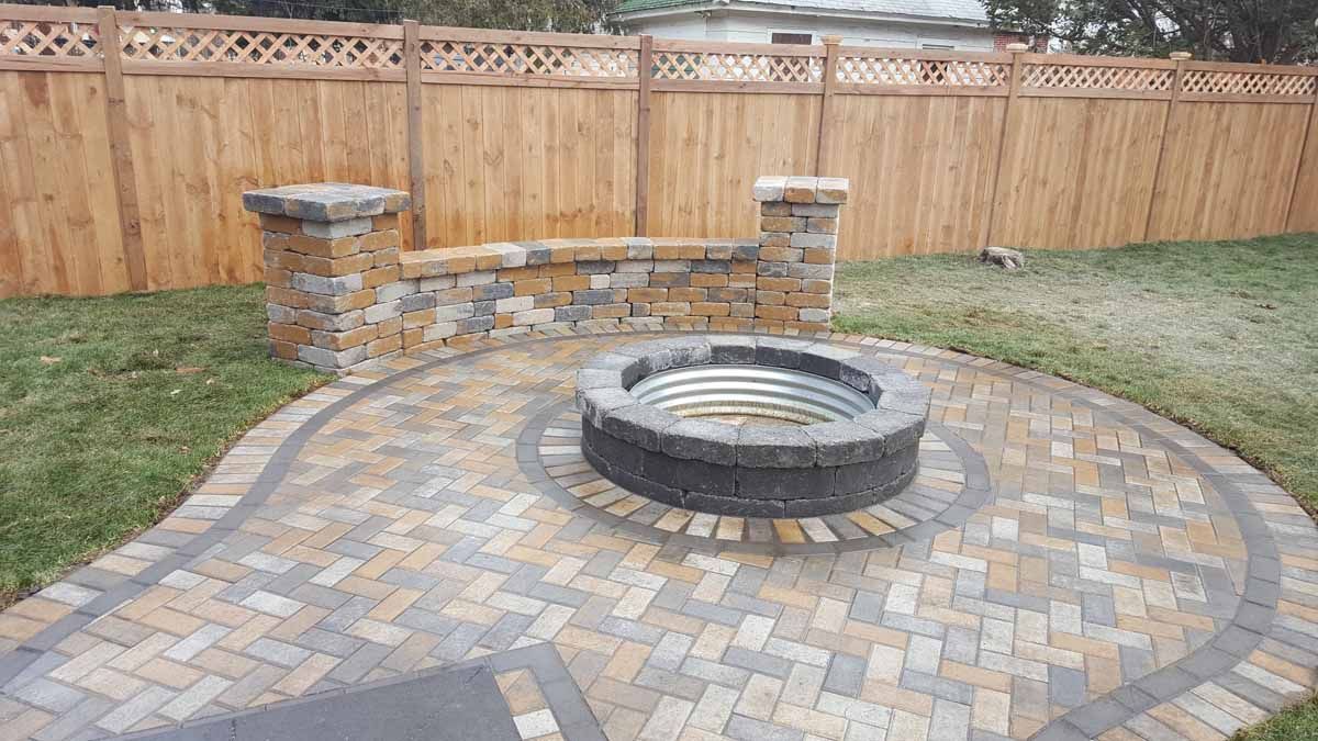 Backyard stone fire pit surrounded by a paver patio and wooden fence