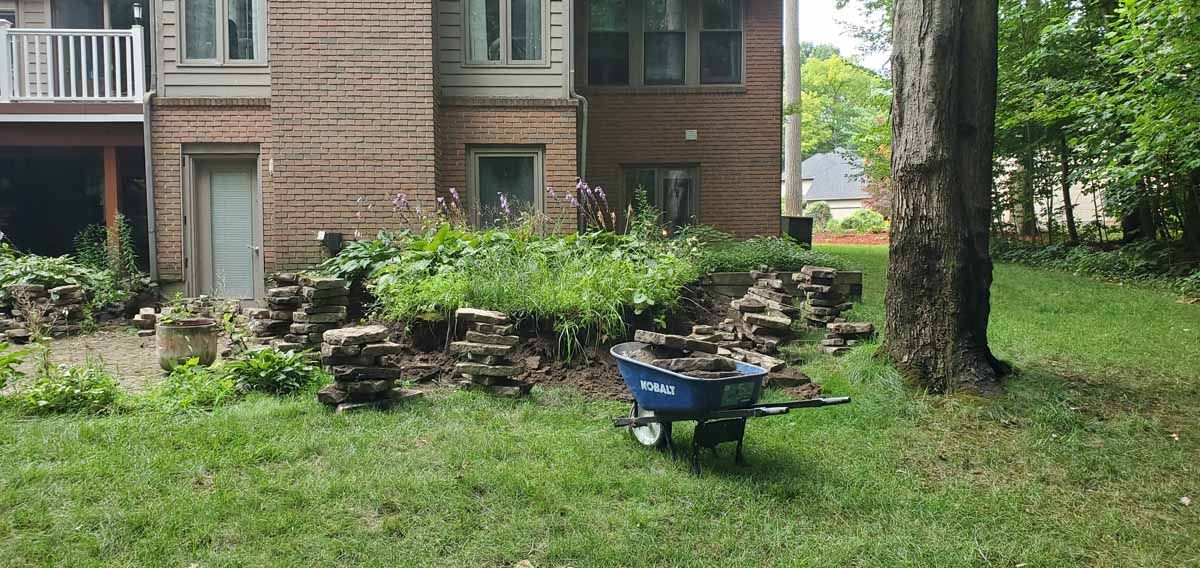 Brick apartment with small garden, lawn, trees, stone steps, and a blue wheelbarrow in the foreground