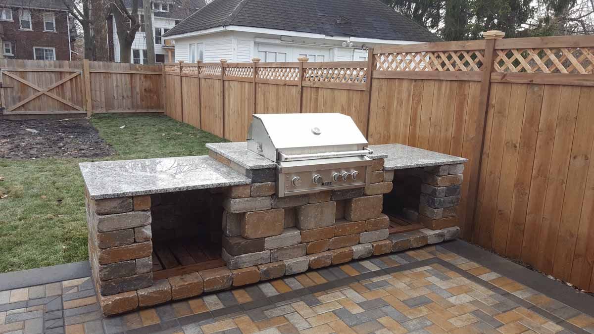 Outdoor stone-and-brick grill with metal lid in a fenced backyard patio