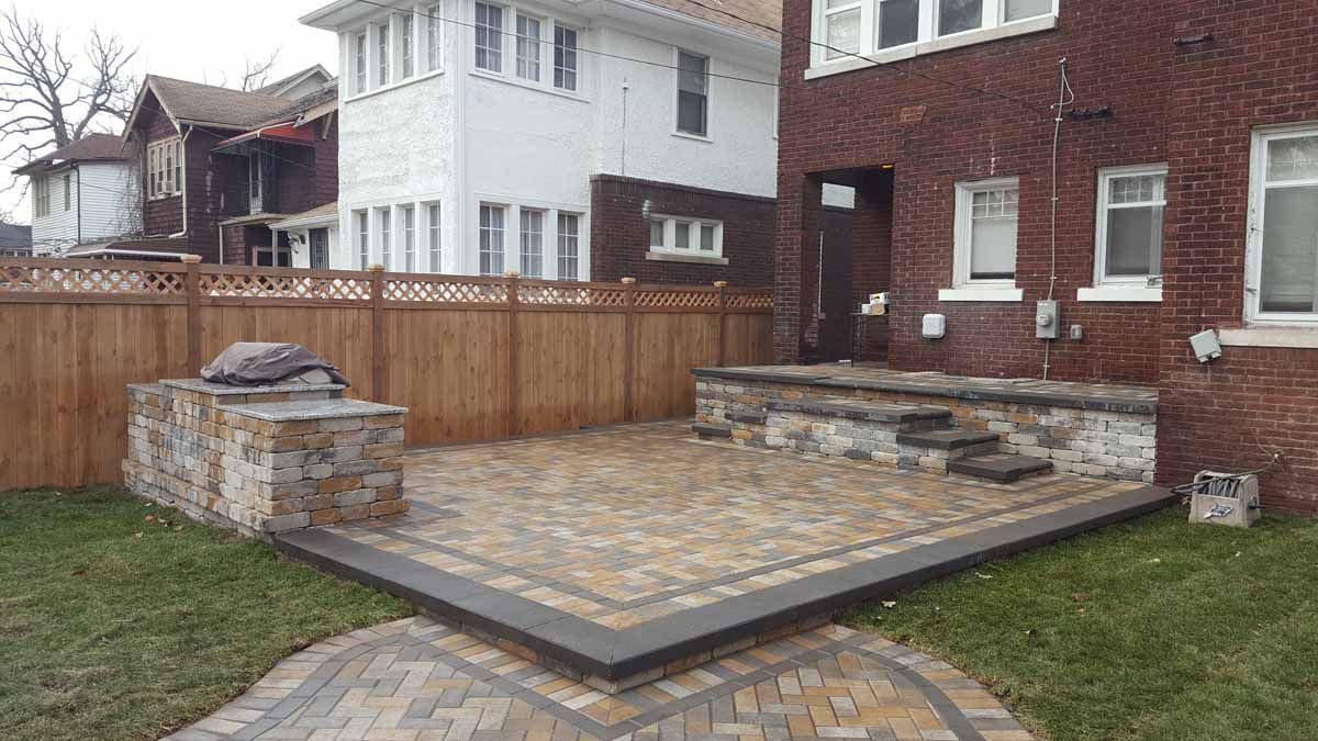 Backyard patio with stone pavers, a low wooden fence, and brick and white houses in the background