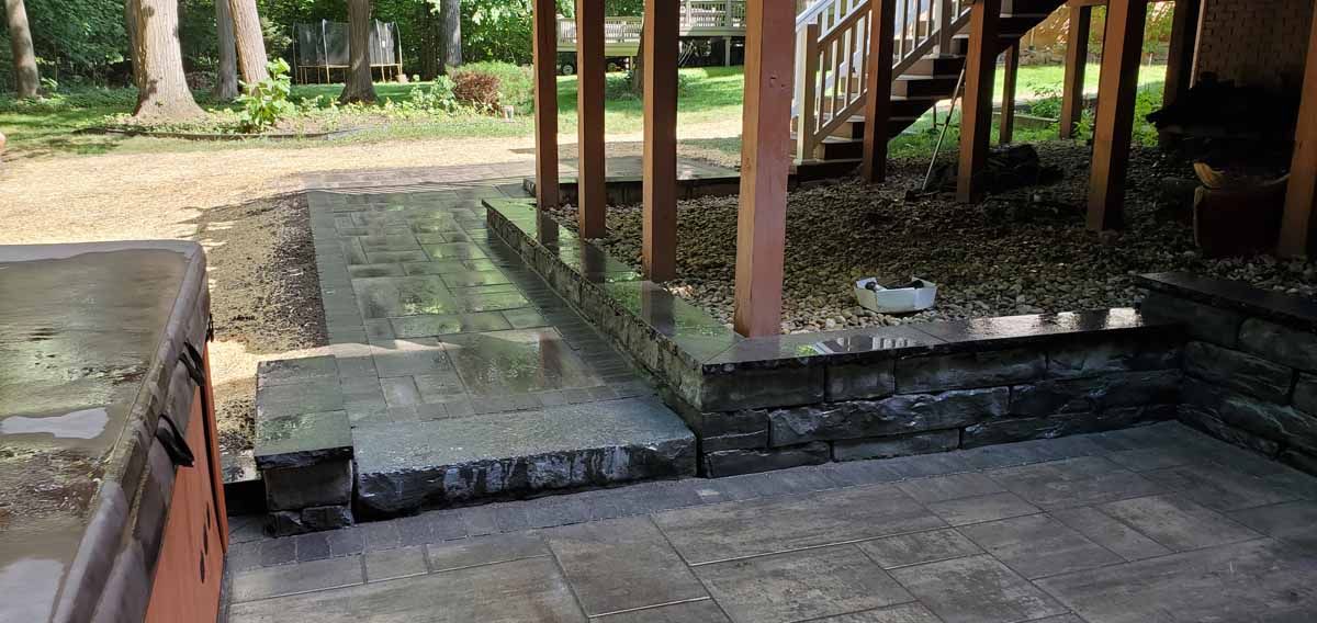 Outdoor stone patio with steps, a wooden deck, and a low retaining wall beside a grassy area