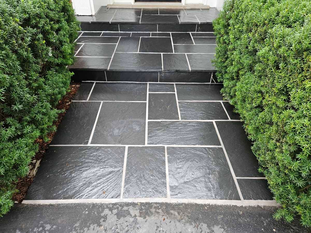 Stone pathway with gray tiles and green shrubs on both sides leading to steps.