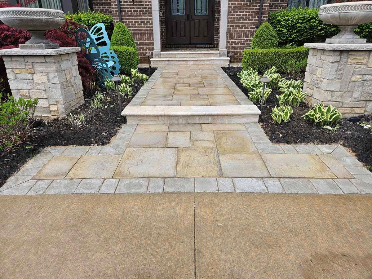 Front walkway with stone pavers leading to a house entrance, bordered by landscaping and stone pillars.