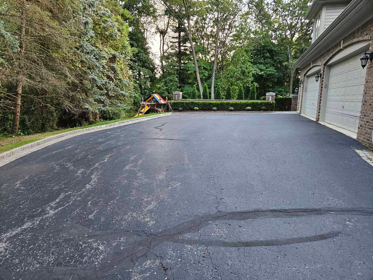 Fresh black asphalt driveway beside a garage, with trees and a small play area at the end.