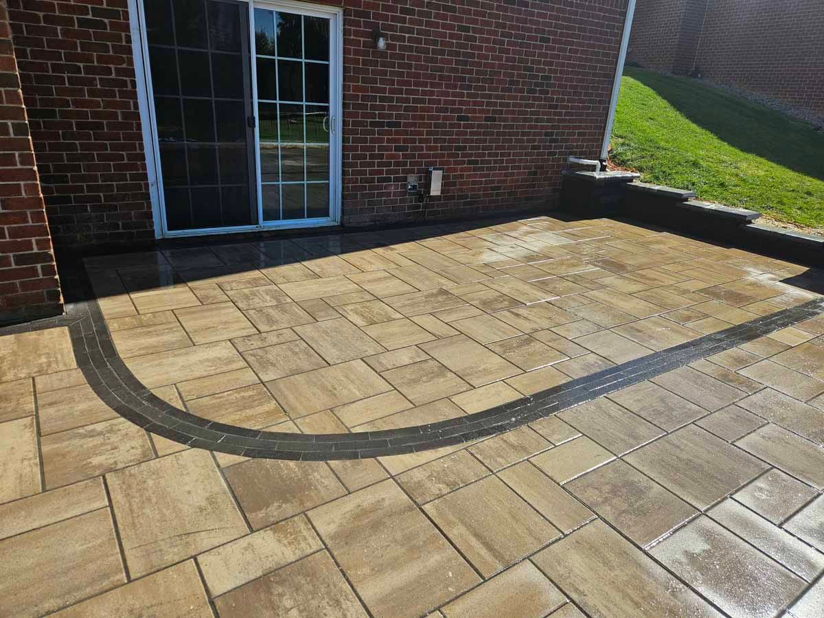 Brick patio with curved dark border beside a house and sliding glass door in sunlight
