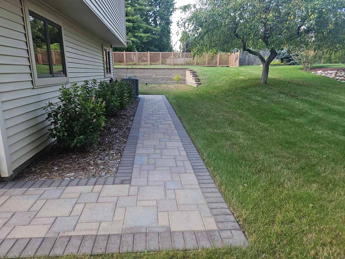 Brick paver walkway along a house, bordered by shrubs and grass, leading to a wooden fence.
