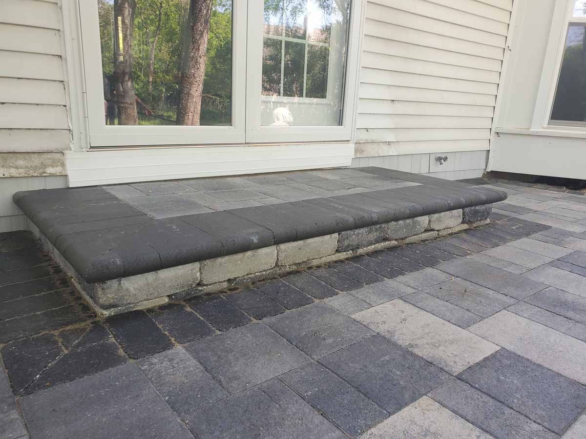 Black-cushioned porch bench beside a house window on a gray paver patio