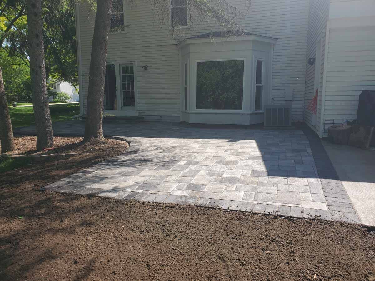 Front yard with a newly paved gray stone patio beside a white house, bordered by dirt and trees